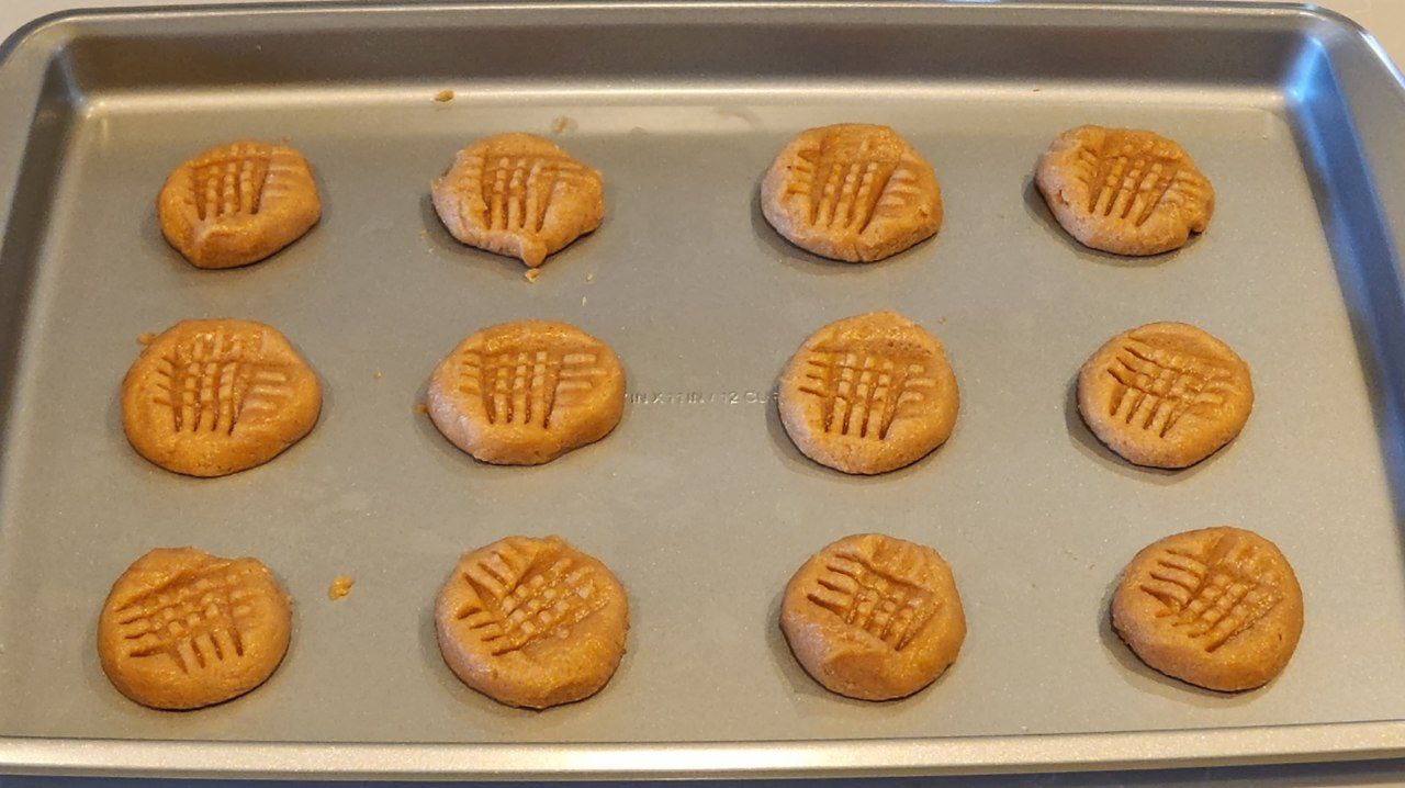 twelve raw cookies on a baking sheet with crosshatch patterns made by a fork